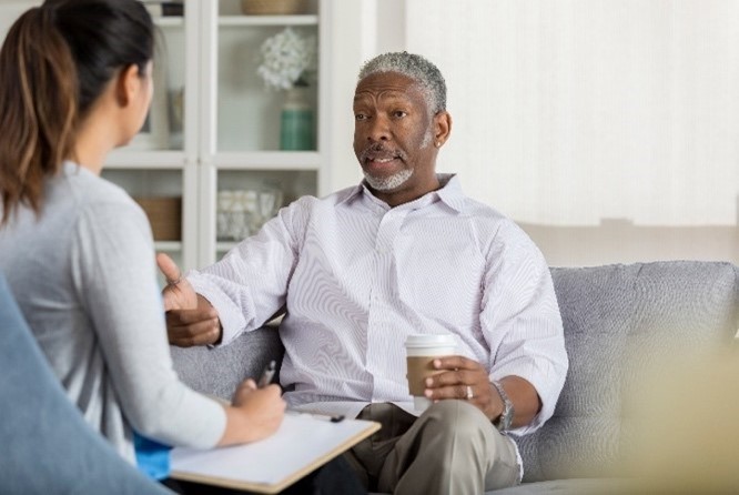 Elderly black man in counselling, sitting on a couch and talking to a woman who has their back to the camera.